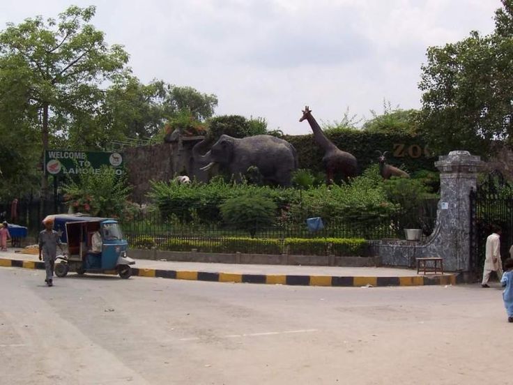 Main entrance of Lahore Zoo with animal sculptures, a rickshaw, and pedestrians near the entrance gate.